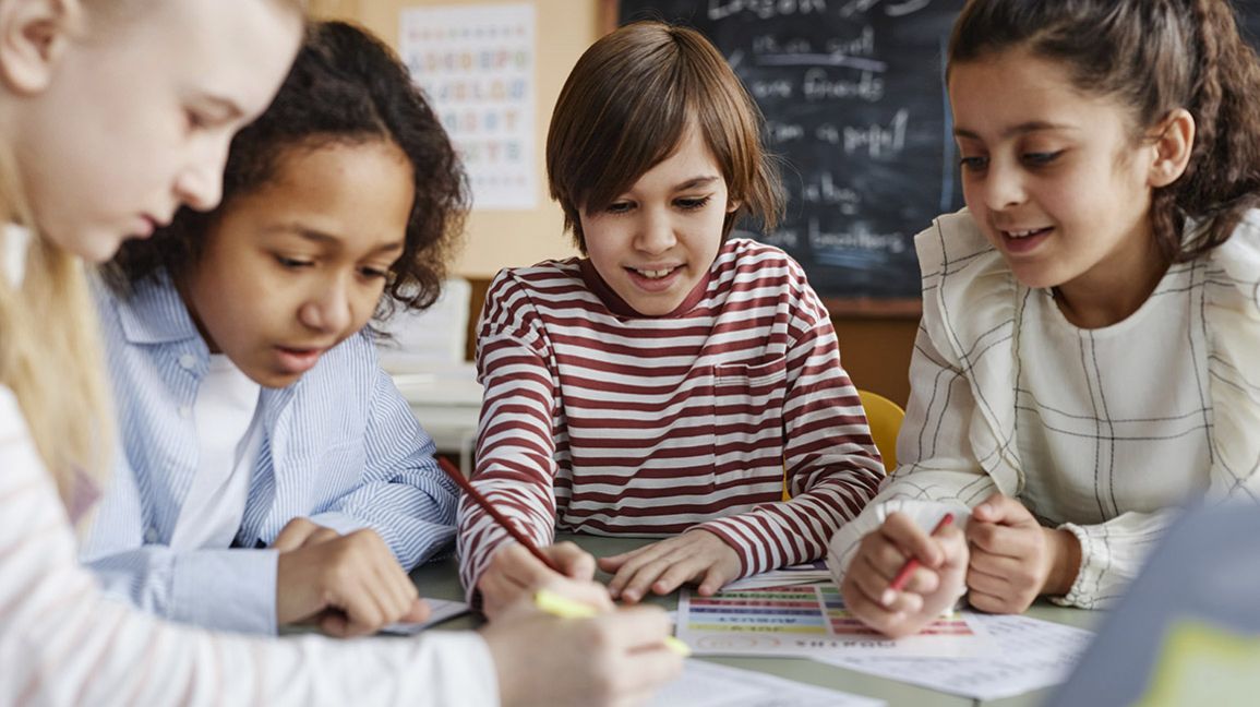 Eine Gruppe ethnisch gemischter Kinder sitzt an einem Tisch im Klassenzimmer und macht gemeinsam Übungen wäh-rend des Unterrichts.