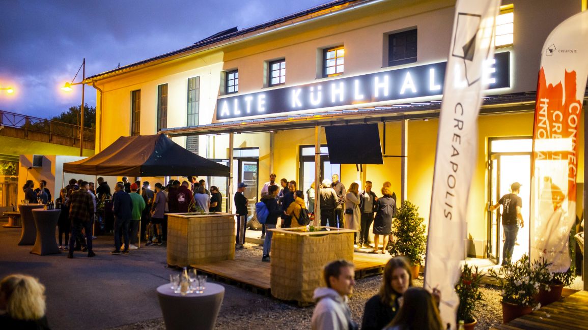 Eine große Gruppe von Personen steht vor einem erleuchteten Haus mit Stehtischen und einem Pavillon davor.